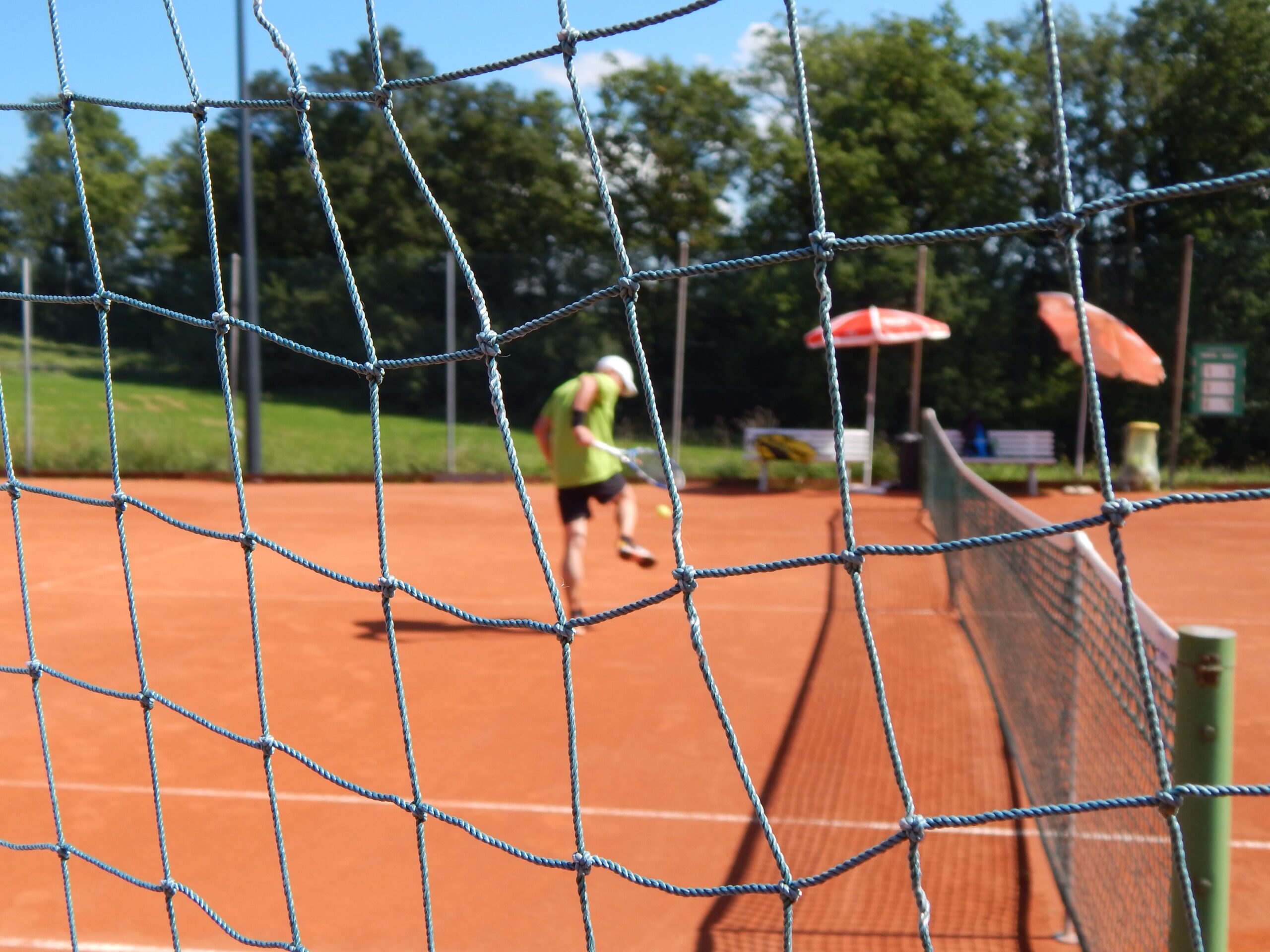 Der Blick durch ein Netz auf einen Tennisplatz, wo eine Person einen Tennisball aufhebt.