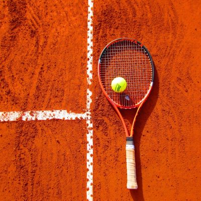 tennis-1671852_1920 Ein Tennisbal liegt auf einem Tennisschläger auf einem Sandplatz.